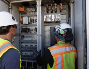 Two workers wearing hard hats and safety vests inspect an open electrical panel filled with various components and wiring.