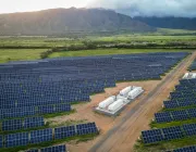 Aerial view of a large solar farm with rows of solar panels on green fields. Several white utility buildings are visible among the panels. In the background, there are mountains partially covered by clouds.