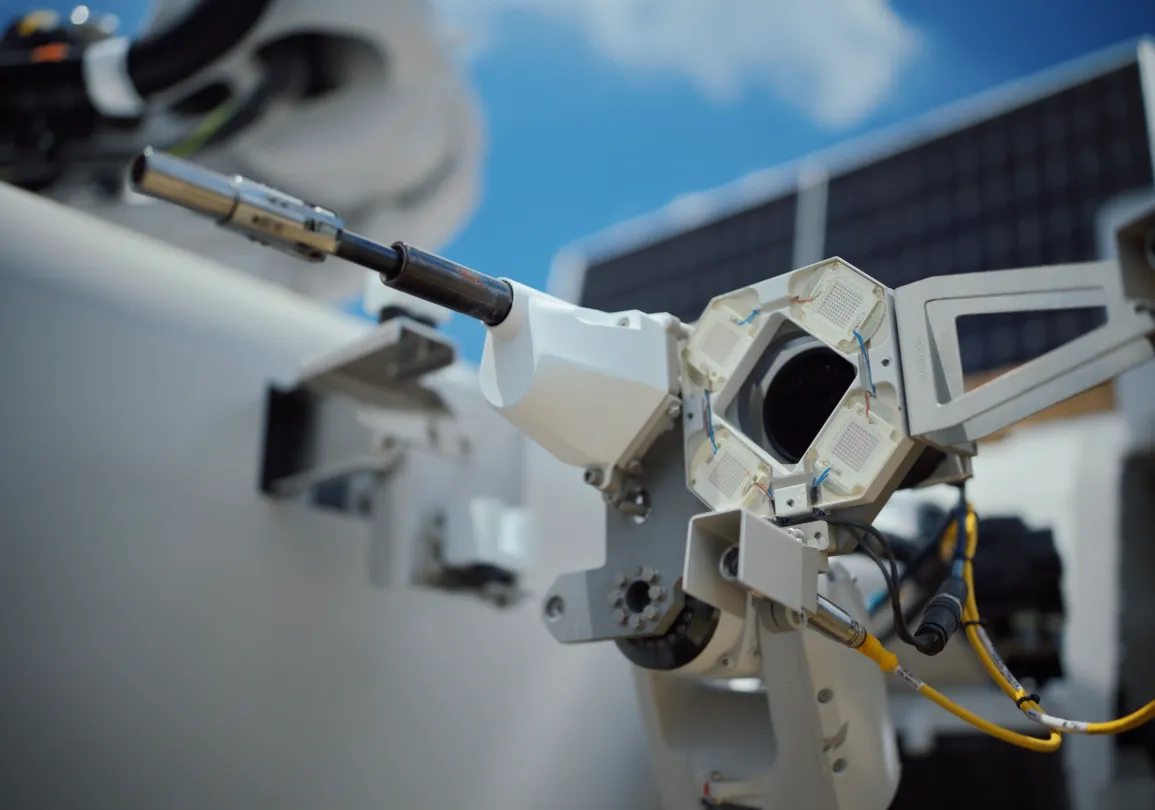Close-up of a robotic arm with a metallic tool, featuring wires and mechanical components, set against a blue sky with a solar panel in the background.