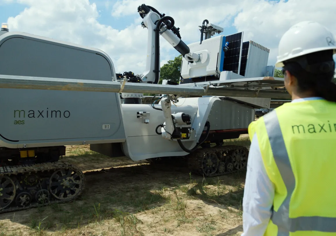 A person wearing a hard hat and a high-visibility vest with 'maximo' on it stands near a large robotic machine on tracks, labeled 'maximo aes', in an outdoor setting with a blue sky and clouds.