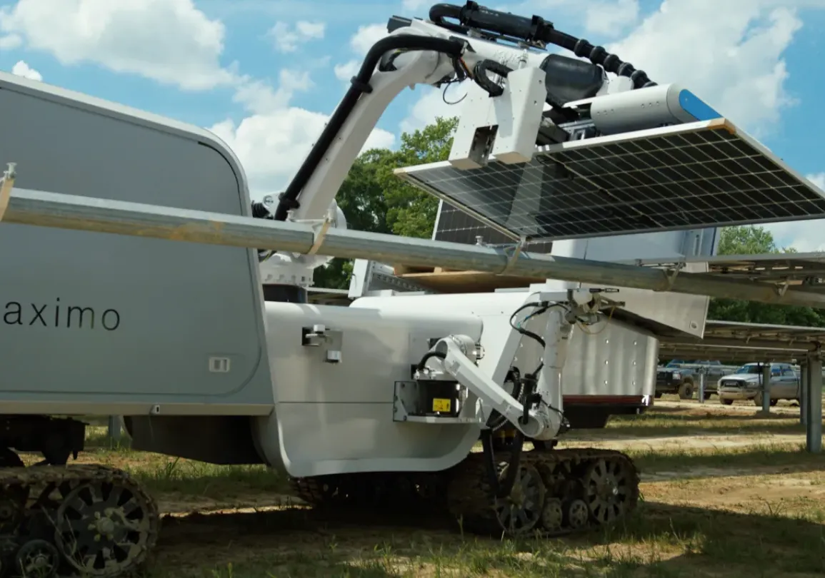 A large robotic machine with tracks is adjusting solar panels in a field. A person wearing a hard hat and reflective vest observes the process. The sky is partly cloudy.