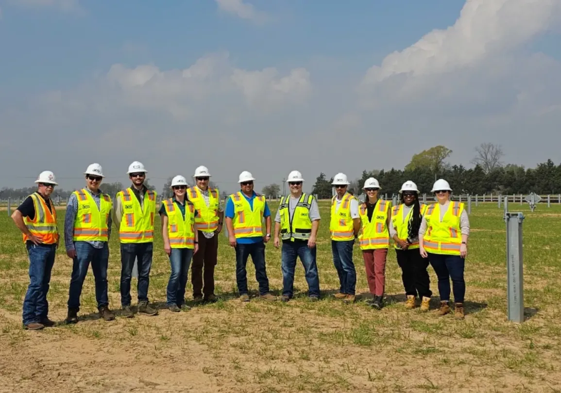 A group of construction workers wearing safety vests and helmets stand in a grassy field with metal posts, under a blue sky with clouds.