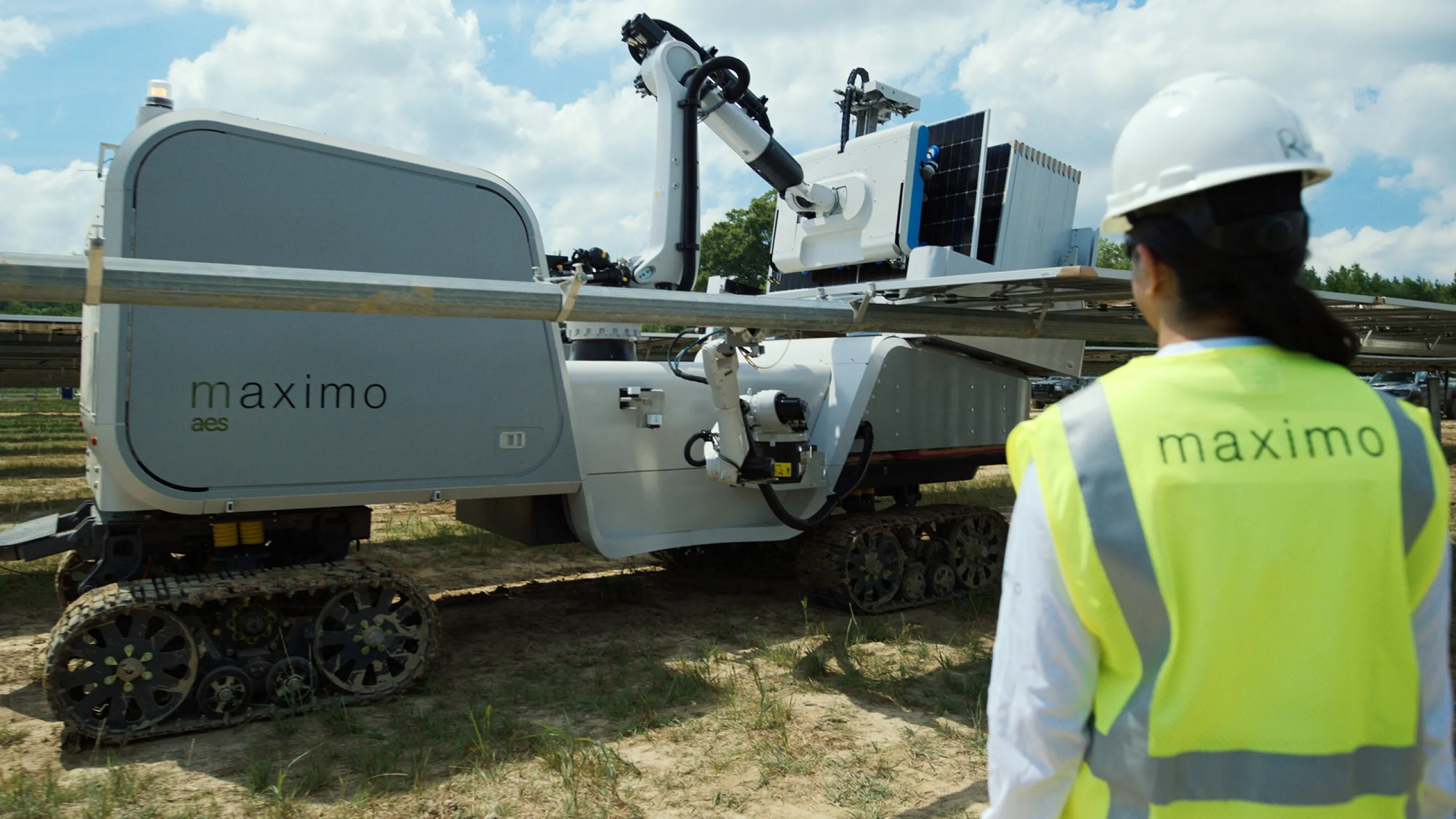 A person wearing a hard hat and a high-visibility vest with 'maximo' on it stands near a large robotic machine on tracks, labeled 'maximo aes', in an outdoor setting with a blue sky and clouds.